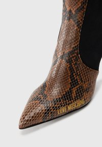 Spisse tå brune og svarte støvler i slangeskinnmønster med svart elastisk sidepanel og "LOVE MOSCHINO" i gullbokstaver nær tåen.