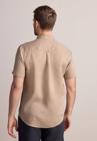 Beiges Leinenhemd mit kurzen Ärmeln, Knopfleiste am Kragen, Rückenfalte und geschwungenem Saum. Kombiniert mit dunkler Hose.