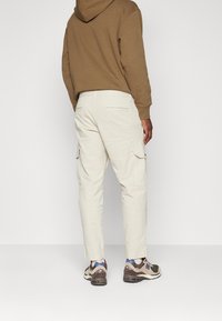 Béžové kalhoty typu cargo s bočními kapsami, rovný střih nohavic a hladkou texturou, doplněné o vícebarevné tenisky a hnědou mikinu s kapucí.
