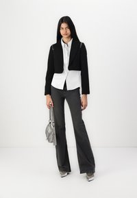 Svart kort blazer med knappdetaljer på skuldrene, hvit skjorte med knapper, mørke slengjeans og en sølvfarget håndveske med stropp.