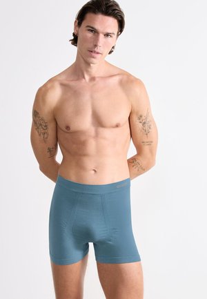 Oberkörperfreier Mann mit mittellangen Haaren, der türkisfarbene Boxershorts trägt, vor einem schlichten weißen Hintergrund steht und Tätowierungen auf beiden Armen hat.