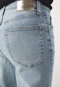 Jean en denim bleu clair avec surpiqûres sur la poche arrière, un petit logo brodé et une pièce métallique texturée sur la ceinture.