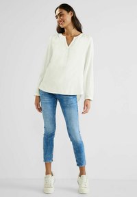 Camicia bianca a maniche lunghe con scollatura a V e dettagli in bottone, abbinata a jeans skinny blu chiaro e sneakers bianche. Tessuto morbido.