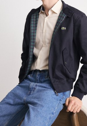 Marineblaue Reißverschlussjacke mit grünem Karofutter, kragenförmigem Ausschnitt, zwei Vordertaschen und einem Lacoste-Logo. Kombiniert mit hellblauen Jeans.