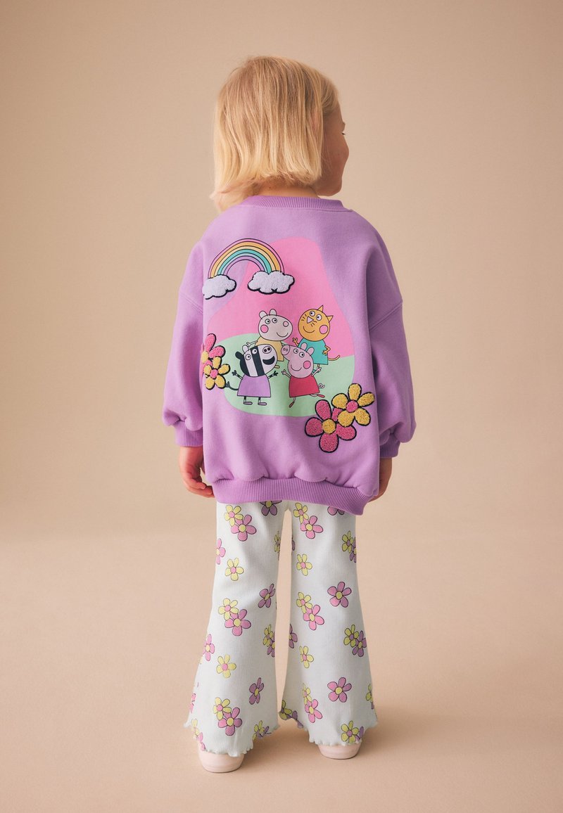 Niño con sudadera lavanda con animales de dibujos animados y flores, combinada con pantalones blancos decorados con flores rosas y amarillas, de pie de espaldas.