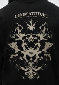 Sweat à capuche noir avec un motif floral ornemental beige et le texte "Denim Attitude Jack and Jones" dans le dos.