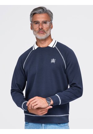 Mand med gråt hår og skæg iført klare briller, en marineblå sweatshirt med hvid krave og emblem, blå jeans og et sølvfarvet armbåndsur.