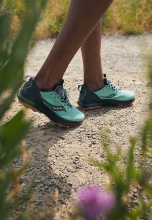 Personne portant des chaussures de course sur sentier de couleur bleu sarcelle et noir, marchant sur un chemin de gravier entouré de verdure.