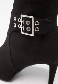 Bottine noire en daim avec un haut talon aiguille. Présente une boucle en argent et des œillets métalliques sur une sangle décorative. Texture lisse.