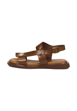 Sandale plate en cuir marron avec bride de cheville réglable, deux sangles horizontales sur le pied et un médaillon décoratif en métal rond sur la bride avant.