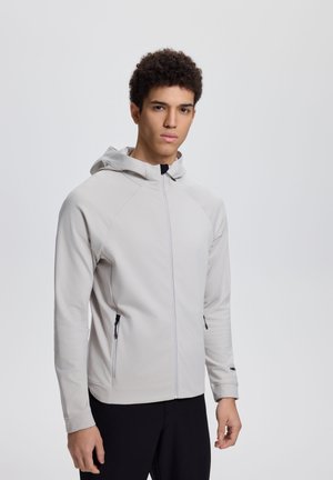 Nuori mies, joka pitää päällään vaaleanharmaata vetoketjullista hupparia ja mustia housuja, seisoo yksiväristä vaaleaa taustaa vasten.