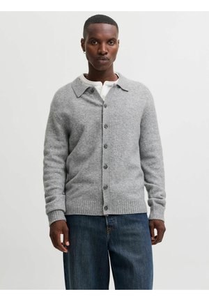 Homme portant un cardigan gris clair boutonné par-dessus une chemise blanche et un jean bleu, debout devant un fond clair uni.
