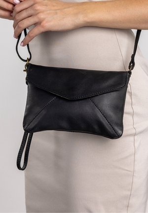 Main tenant un sac bandoulière en cuir noir style enveloppe porté sur une tenue beige avec bracelet au poignet et fermeture à glissière.