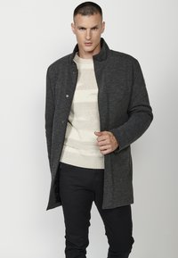 Chaqueta exterior de un gris oscuro con un alto cuello, cierres a presión y mangas largas, superpuesta a un suéter de punto texturizado en color crema.