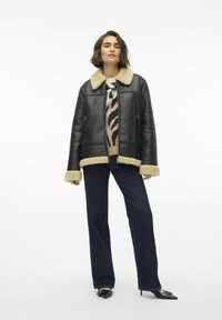 Veste en cuir noire avec col et poignets en fourrure beige, associée à un pull à motifs, un jean bleu foncé et des chaussures noires à bouts pointus.