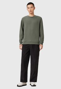 Sudadera verde de tela suave con cuello redondo, combinada con pantalones de pana negros sueltos que tienen dos bolsillos frontales y un acabado texturizado.