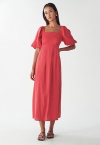 Rdeča midi obleka s kvadratnim izrezom, napihnjenimi rokavi in nabranim dizajnom. Gladka tekstura, tekoča tkanina, oblečena z črnimi sandali s trakovi.