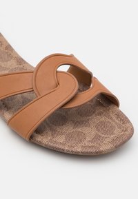 Brune faux læder sandaler med et buet stropdesign, tekstureret fodseng med et geometrisk mønster og en kontureret form.