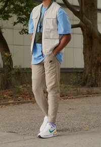 Beiga fleecebyxor med struktur, ljusblå t-shirt, cremefärgad nyckelväst med fickor och vita Nike-sneakers med gröna och gula detaljer.