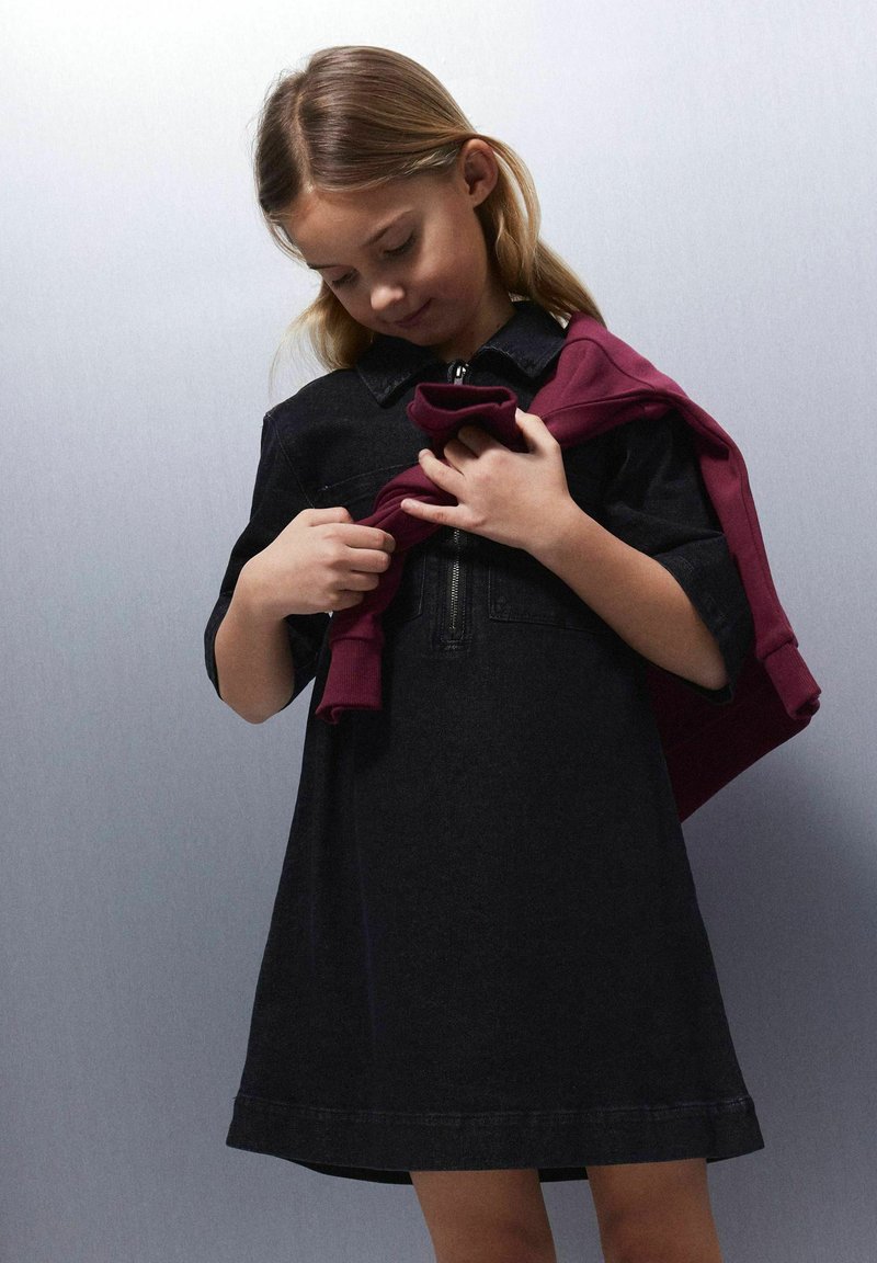 Robe en denim bleu indigo foncé avec un col et une fermeture éclair sur le devant, portée avec un sweat-shirt bordeaux drapé sur un bras. Texture de tissu lisse.