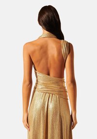 Robe dorée sans dos avec un design drapé, présentant un buste enveloppé. Le tissu a une texture scintillante et descend en une jupe évasée.