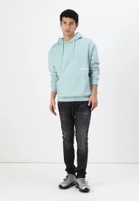 Svetlo modra kapuca s sprednjim žepom in vrvicami, v kombinaciji s temnimi denim hlačami in sivimi športnimi čevlji. Minimalno vidno blagovno znamko.