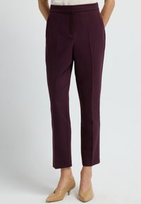 Vrouw in op maat gemaakte bordeauxrode broek tot de enkel en beige lage puntige schoenen, staand tegen een effen achtergrond.