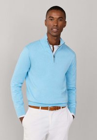 Mężczyzna w jasnoniebieskim swetrze z krótkim zamkiem na 1/4 długości, nałożonym na białą koszulę, białe spodnie i brązowy pasek, stoi z rękami w kieszeniach.