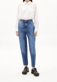 Woman wearing high-waist blue jeans, a white button-up shirt tucked in, and black platform clogs standing against a plain white background.