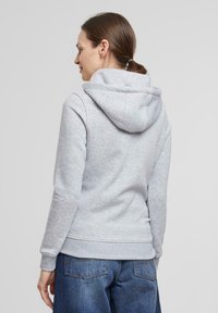 Sudadera gris con capucha ajustable, puños y dobladillo acanalados. Textura suave y corte casual, combinada con jeans de mezclilla oscuros. Vista trasera.