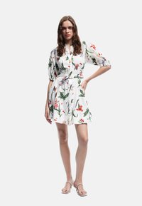 Bloemen shirtjurk van witte stof met kleurrijke patronen van bloemen en bladeren, met poffende mouwen en een tailleband.