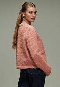 Pink fuzzy sweater with a rounded neckline, featuring a decorative beaded collar and side pockets, paired with dark denim jeans.