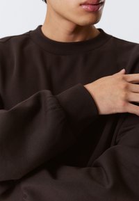 Gros plan sur une personne portant un sweat-shirt à manches longues marron foncé avec des poignets côtelés, reposant sa main sur l'épaule opposée, lèvres et menton visibles.