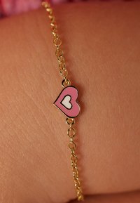 Gouden kettingarmband met een roze hartbedel met een witte binnenhartdetail, rustend op een huidkleurig oppervlak.