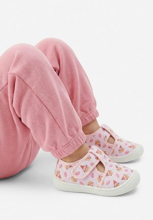 Dziecko w różowych spodniach i różowych trampkach z nadrukami cukierków i babeczek z kreskówek, siedzące z podkurczonymi kolanami na białym tle.