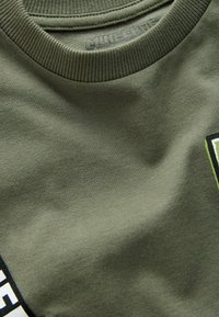 Olivegrünes Baumwoll-T-Shirt mit geripptem Rundhalsausschnitt und kontrastierender Grafik in Schwarz und leuchtendem Grün nahe dem unteren Saum.
