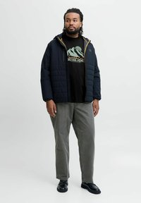 Sort pufferjakke med hætte, med et quiltet mønster og gule accenter. Båret over en grafisk sort t-shirt og grå bukser. Sorte sko.