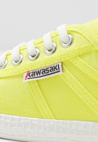 Zapatilla de lona amarillo lima con cordones blancos, ojetes plateados, suela blanca texturizada y etiqueta con el logo "Kawasaki" en rojo y negro.