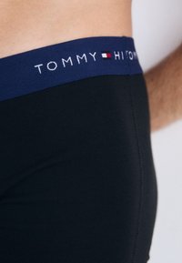 Close-up of a person wearing black underwear with a navy waistband featuring the Tommy Hilfiger logo and flag emblem.