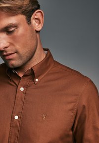 Camisa marrón de botones hecha de tela texturizada, que presenta un pequeño logo bordado y un patrón de cuadros en el interior del cuello.