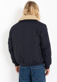 Chaqueta bomber de color marino con un cuello de piel sintética beige, textura acolchada, puños y dobladillo elásticos. Presenta un corte relajado y bolsillos laterales.