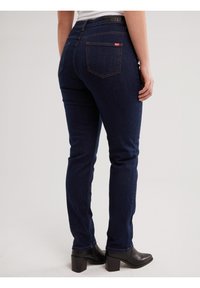 Persona valkā tumši zilas pieguļošas džinsu bikses ar aizmugures kabatām un melnus potīšu zābakus ar papēdi, stāvot pret vienkāršu gaišu fona sienu.