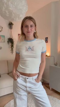 Top corto bianco con rifinitura azzurro chiaro, caratterizzato da una grafica di un cartone animato che raffigura una creatura blu con ali. Indossato con jeans bianchi a gamba larga.
