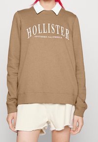 Brun sweatshirt med vit "HOLLISTER" text, ribbade ärmslut och fåll, över en vit skjorta med krage. Bärs med fransiga vita shorts.