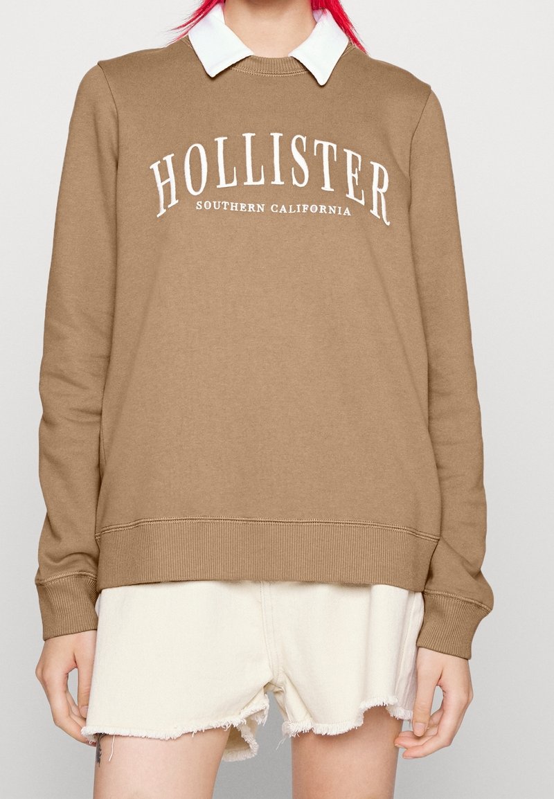 Brun sweatshirt med vit "HOLLISTER" text, ribbade ärmslut och fåll, över en vit skjorta med krage. Bärs med fransiga vita shorts.