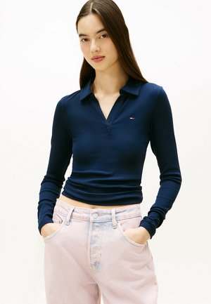 Mujer joven con cabello largo y oscuro, lleva una camiseta de manga larga azul marino con textura acanalada y pantalones vaqueros de tiro alto color rosa claro, de pie con las manos en los bolsillos.