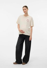 Zwangere vrouw in een beige t-shirt en brede zwarte broek, zachtjes haar buik vasthoudend, staand tegen een effen witte achtergrond.