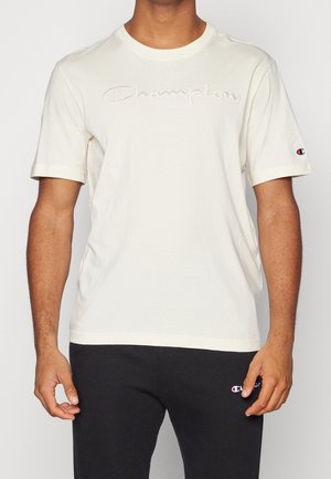 Mann trägt ein cremefarbenes Champion-T-Shirt mit besticktem Logo und schwarze Champion-Sweathose.