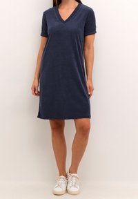 Robe bleu marine à manches courtes et col en V en tissu doux, longueur genou avec manches retroussées, portée avec des baskets blanches. Design simple et décontracté.