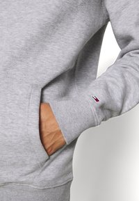 Szara bluza z prążkowanymi mankietami i dolnym ściągaczem, z boczną kieszenią oraz małą naszywką z logo w kolorach czerwonym, białym i niebieskim na rękawie.